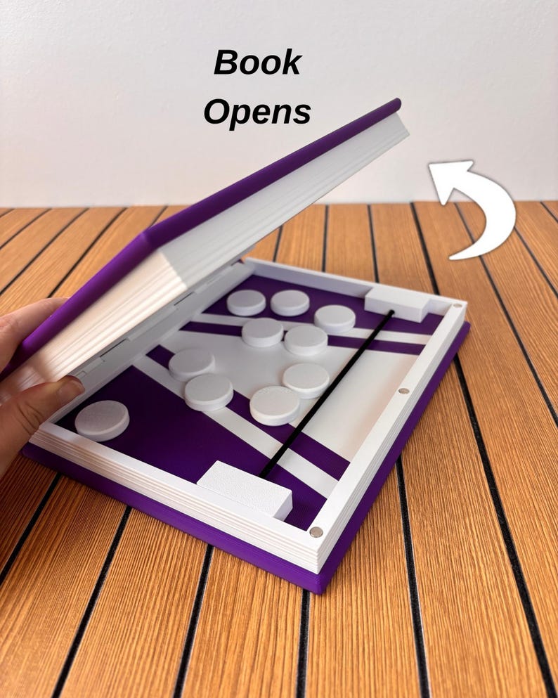 May include: A purple and white game board shaped like a book with the text Book Opens. The interior features a purple surface with white lines, white circular game pieces, and a black elastic band. A white arrow indicates the opening direction.