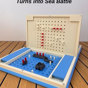 May include: A board game featuring a blue grid base with gray plastic ships and red pegs, and a cream-colored vertical grid board with rows A-J and columns 1-10. The text 'Turns into Sea Battle' is printed above the game board in black font.