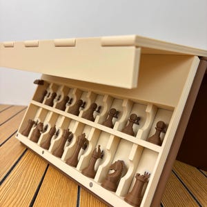 K&ouml;nnte beinhalten: Ein beigefarbenes und braunes, zusammenklappbares Schachspiel-Etui mit ge&ouml;ffnetem Deckel, das zwei innere Reihen geformter F&auml;cher enth&uuml;llt. Jedes Fach enth&auml;lt eine braune Schachfigur, darunter Bauern, Springer, Turm, L&auml;ufer und K&ouml;nig.