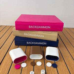 Könnte beinhalten: Ein Stapel aus drei rechteckigen Backgammon-Koffern in Pink, Beige und Marineblau, jeweils mit der Aufschrift BACKGAMMON. Darunter befinden sich drei ovale Würfelbecher in Weiß, Braun und Weiß sowie sechs kleine runde Spielsteine und zwei weiße Würfel.