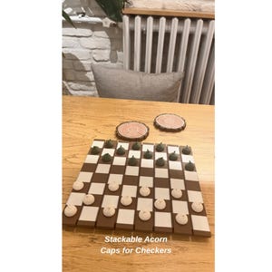 K&ouml;nnte beinhalten: Ein Schachbrett mit braunen und cremefarbenen Feldern, mit eichelf&ouml;rmigen Spielsteinen in Gr&uuml;n und Creme. Zwei Holzuntersetzer mit Baumring-Designs sind ebenfalls sichtbar. Der Text "Stackable Acorn Caps for Checkers" befindet sich unten.