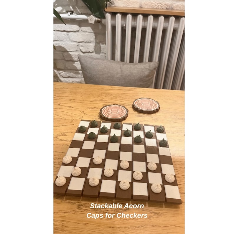 K&ouml;nnte beinhalten: Ein Schachbrett mit braunen und cremefarbenen Feldern, mit eichelf&ouml;rmigen Spielsteinen in Gr&uuml;n und Creme. Zwei Holzuntersetzer mit Baumring-Designs sind ebenfalls sichtbar. Der Text "Stackable Acorn Caps for Checkers" befindet sich unten.