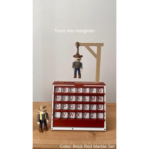 May include: A brick red and white Hangman game set with a cowboy figure. The game features a gallows with a hanging cowboy figure and a letter board. The text "Turns Into Hangman" is visible. The colour is described as Brick Red Marble Set.