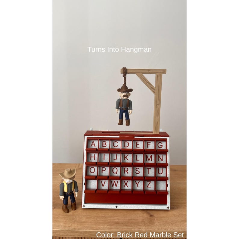 May include: A brick red and white Hangman game set with a cowboy figure. The game features a gallows with a hanging cowboy figure and a letter board. The text "Turns Into Hangman" is visible. The colour is described as Brick Red Marble Set.