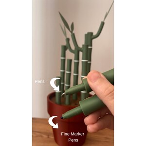 May include: A close-up of two green fine marker pens, one with a white tip and the other with a blue tip, held in a hand. The pens are labeled "Fine Marker Pens". A decorative bamboo plant in a brown pot is in the background.