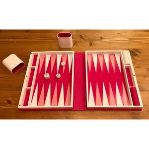 May include: A backgammon set with a vibrant pink and white color scheme. The game board is open, revealing the playing surface with alternating pink and white triangles. Dice and game pieces are visible, along with two white and pink containers.