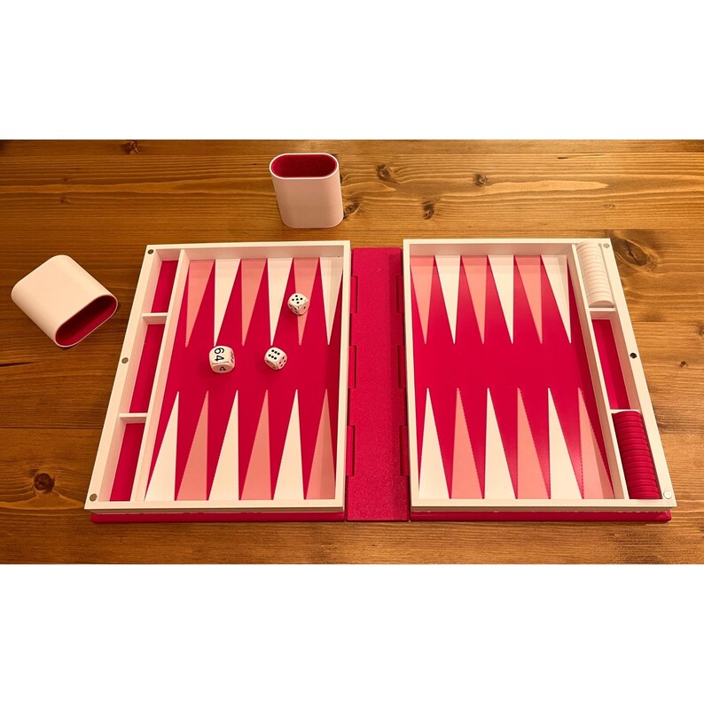 May include: A backgammon set with a vibrant pink and white color scheme. The game board is open, revealing the playing surface with alternating pink and white triangles. Dice and game pieces are visible, along with two white and pink containers.