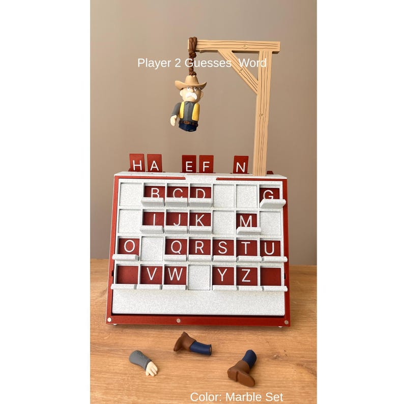 May include: A red and white word guessing game with a gallows-style structure and a small figure. The game board has letter tiles and the text "Player 2 Guesses Word." The color is described as "Marble Set."