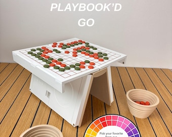 May include: A white board game table labelled PLAYBOOK'D GO with a grid surface featuring orange and green circular game pieces. Two beige ribbed bowls hold additional pieces. A circular colour wheel graphic displays 33 unique colour choices.