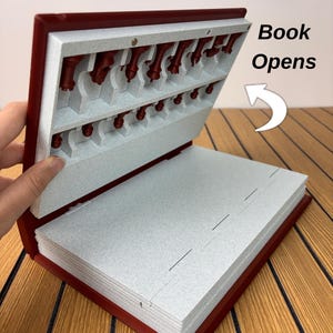 K&ouml;nnte beinhalten: Ein rotes, buchf&ouml;rmiges Aufbewahrungsetui mit gesprenkeltem grauen Innenraum und geformten F&auml;chern f&uuml;r Schachfiguren. Der Deckel ist teilweise ge&ouml;ffnet und gibt den Blick auf die Figuren frei. Text "Book Opens" mit einem wei&szlig;en Pfeil.