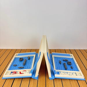May include: A portable battleship board game featuring two cream-colored folding cases with blue grid play areas. Each side includes gray plastic ship pieces, white pegs for tracking shots, and red pegs for hits, arranged on a numbered and lettered grid.
