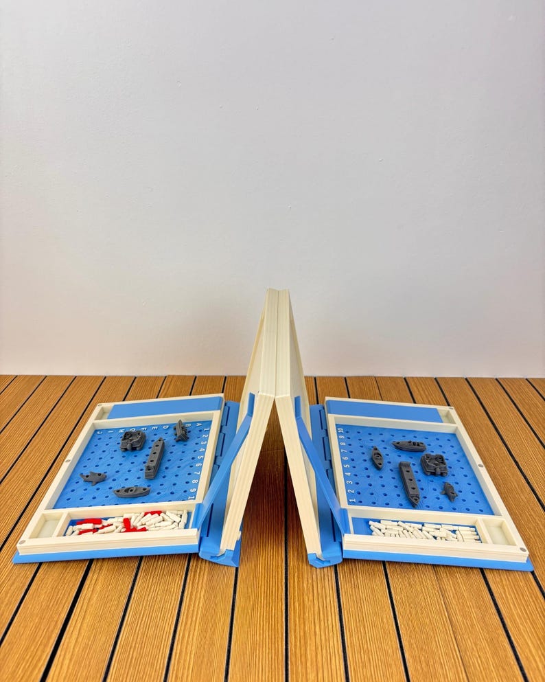 May include: A portable battleship board game featuring two cream-colored folding cases with blue grid play areas. Each side includes gray plastic ship pieces, white pegs for tracking shots, and red pegs for hits, arranged on a numbered and lettered grid.