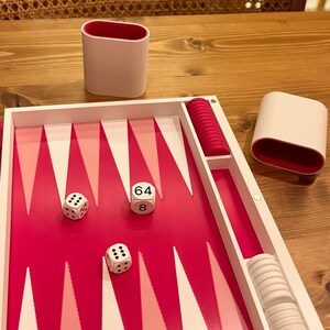 May include: A backgammon set with a white and pink playing surface. The set includes white dice with black dots, white game pieces, and two white and pink dice cups. The dice show the numbers 64 and 8.