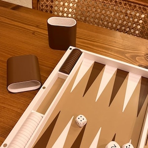 May include: A backgammon set with a brown and white color scheme. The game board features brown and white triangles, dice, and brown playing pieces. Two brown cups with white interiors are also present.