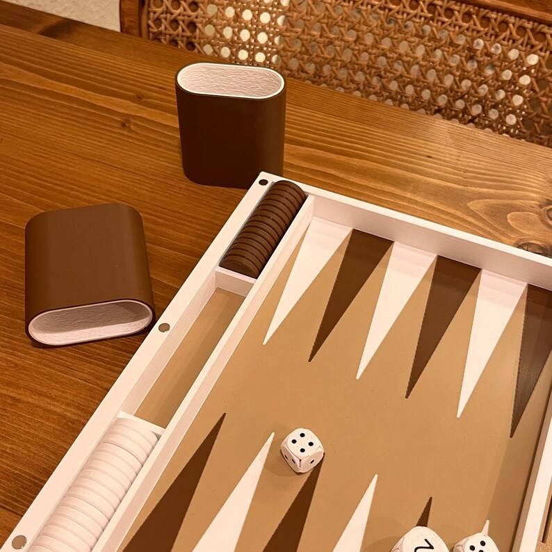 May include: A backgammon set with a brown and white color scheme. The game board features brown and white triangles, dice, and brown playing pieces. Two brown cups with white interiors are also present.