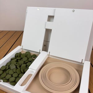 May include: A white open storage organizer featuring a left compartment filled with numerous small, round, olive green discs and a right compartment containing a set of nested, circular, beige silicone rings of varying sizes.