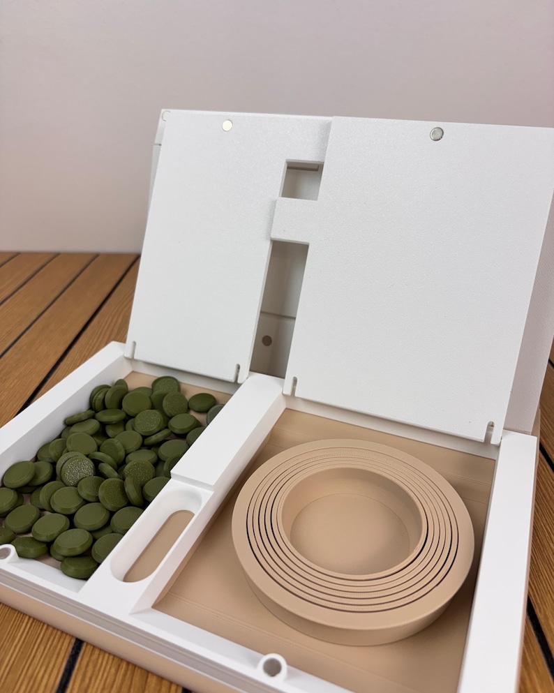 May include: A white open storage organizer featuring a left compartment filled with numerous small, round, olive green discs and a right compartment containing a set of nested, circular, beige silicone rings of varying sizes.