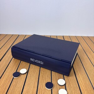 以下が含まれることがあります： ヒンジ付きの蓋が付いた長方形のネイビーブルーのオセロゲームボックス。前面の縁には白い文字で「REVERSI」と印刷されています。ネイビーブルーと白の両面を持つ小さな円形のゲームピースがいくつか散らばっています。