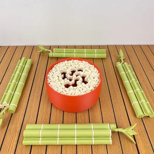 May include: A round, matte red bowl filled with small, white, cylindrical pieces, surrounded by four light green, segmented bamboo-shaped sticks. The bowl features a perforated top surface with a central cutout pattern.