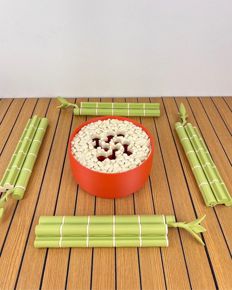 May include: A round, matte red bowl filled with small, white, cylindrical pieces, surrounded by four light green, segmented bamboo-shaped sticks. The bowl features a perforated top surface with a central cutout pattern.