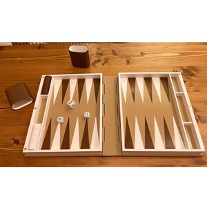 May include: A backgammon set with a brown and white color scheme. The game board is open, displaying the playing surface with brown and white triangles. Dice and game pieces are visible, along with two brown containers. The set is on a wooden surface.