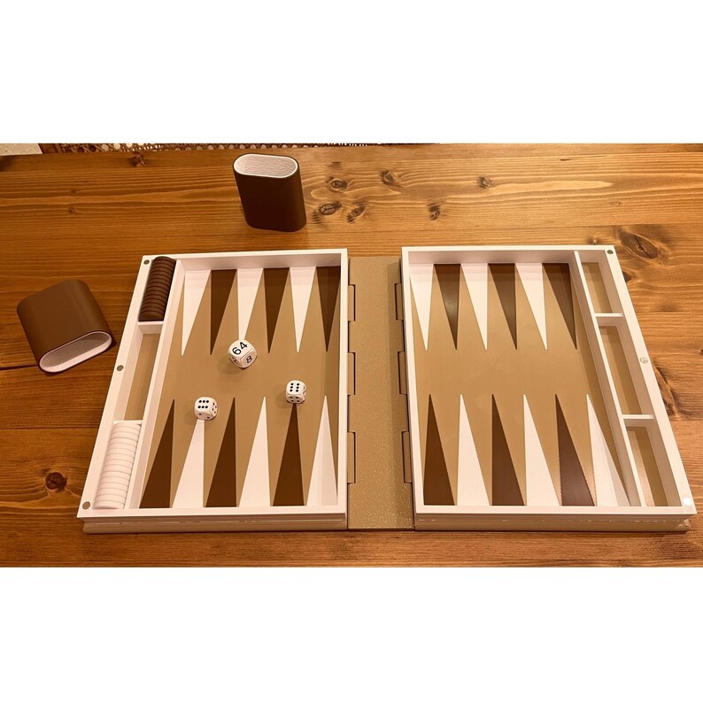 May include: A backgammon set with a brown and white color scheme. The game board is open, displaying the playing surface with brown and white triangles. Dice and game pieces are visible, along with two brown containers. The set is on a wooden surface.