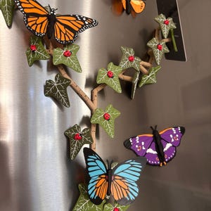 May include: A set of magnetic decorations featuring a tan vine with green ivy leaves, small red ladybugs, and several butterflies in orange, black, blue, and purple colors. The pieces are arranged to look like a climbing plant on a metallic surface.