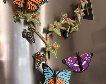 May include: A set of magnetic decorations featuring a tan vine with green ivy leaves, small red ladybugs, and several butterflies in orange, black, blue, and purple colours. The pieces are arranged to look like a climbing plant on a metallic surface.