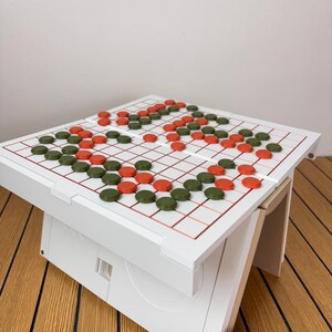 May include: A white board game table featuring a grid with thin red lines. The surface is populated with numerous small, circular game pieces in alternating dark green and orange colors, arranged in various patterns across the playing area.