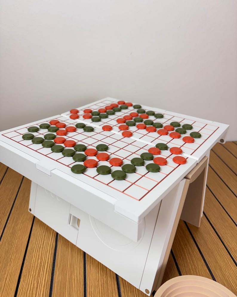 May include: A white board game table featuring a grid with thin red lines. The surface is populated with numerous small, circular game pieces in alternating dark green and orange colors, arranged in various patterns across the playing area.