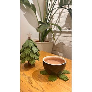May include: A miniature green Christmas tree with a star on top sits next to a black mug filled with a warm beverage. The mug rests on a green leaf-shaped coaster. A wooden table and a potted plant are in the background.