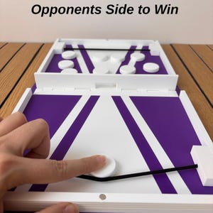 May include: A tabletop board game featuring a purple and white surface with a black elastic band and white circular pucks. The text Get All Pucks On Your Opponents Side to Win is printed above the game board, which includes a central gate.