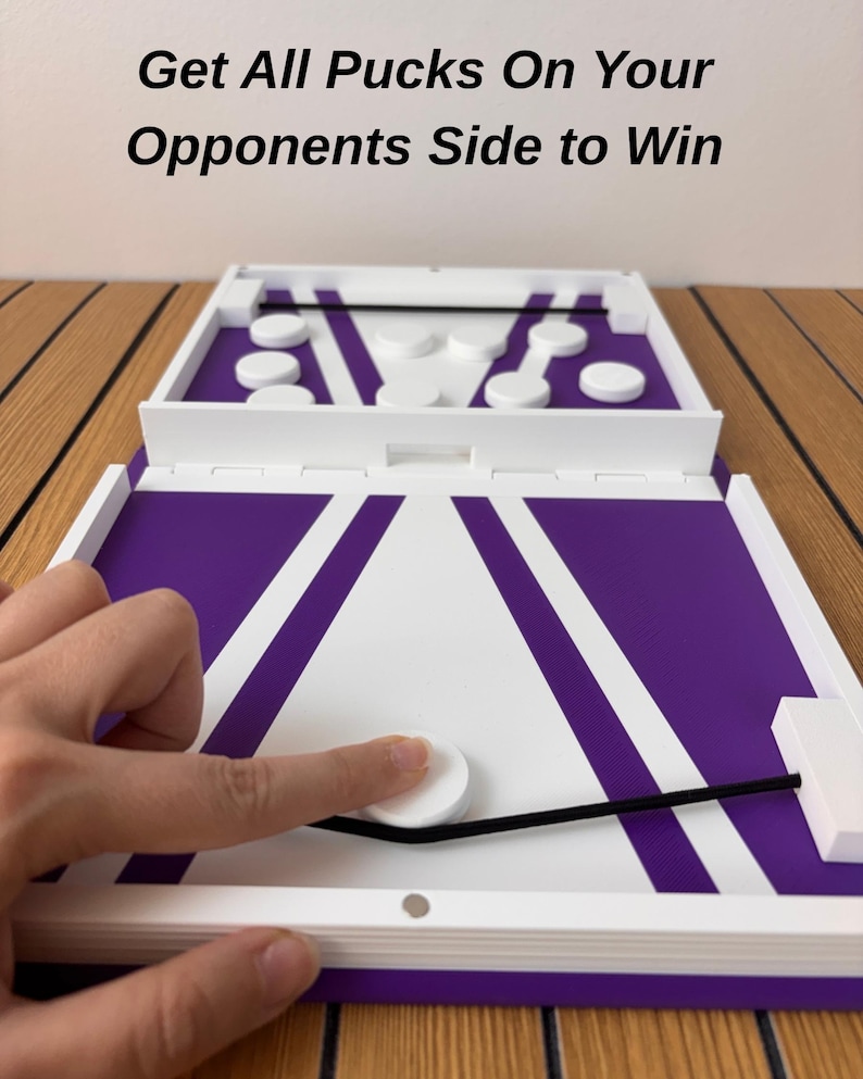 May include: A tabletop board game featuring a purple and white surface with a black elastic band and white circular pucks. The text Get All Pucks On Your Opponents Side to Win is printed above the game board, which includes a central gate.