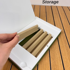 May include: A white and olive green storage case with the text Rack Storage printed on the lid. The interior features a series of tan, angled dividers designed for organizing items, with one divider being lifted by a hand.