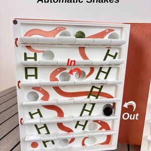 May include: A white board game titled Automatic Snakes featuring orange snake illustrations, green ladder graphics, and circular holes. The board includes red arrows labeled In and Out, with a small green sphere and a black cylinder resting on the shelves.