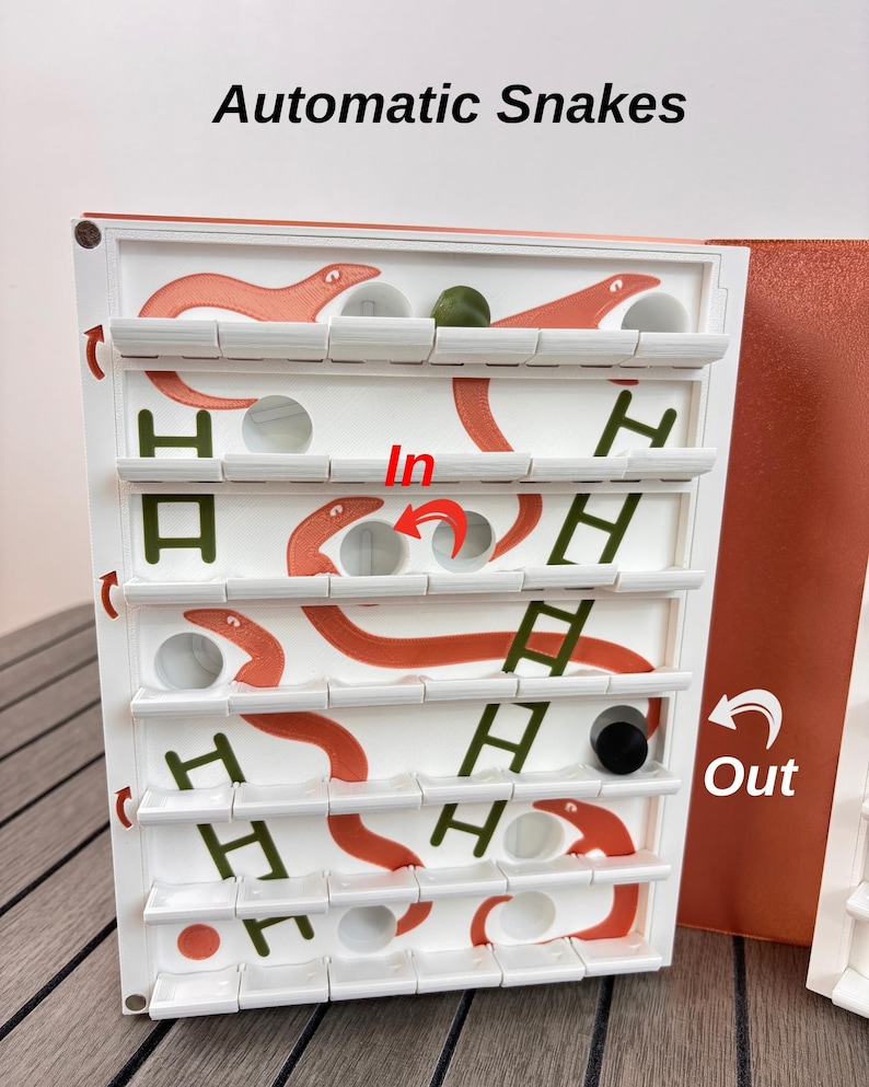 May include: A white board game titled Automatic Snakes featuring orange snake illustrations, green ladder graphics, and circular holes. The board includes red arrows labeled In and Out, with a small green sphere and a black cylinder resting on the shelves.
