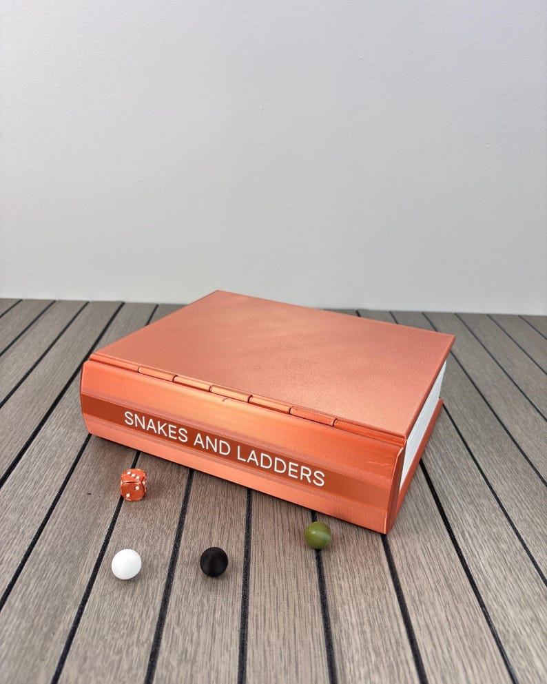 May include: A rectangular, metallic copper-colored board game box with the text "SNAKES AND LADDERS" printed on the spine. The set includes one orange six-sided die, one white marble, one black marble, and one olive green marble.