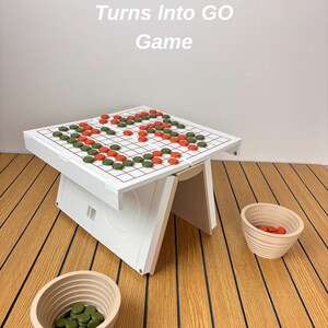 May include: A white Go game board with a red grid, featuring green and orange circular game pieces arranged on the surface. The text "Turns Into GO Game" is visible above the board. Two beige, ribbed bowls containing additional game pieces.