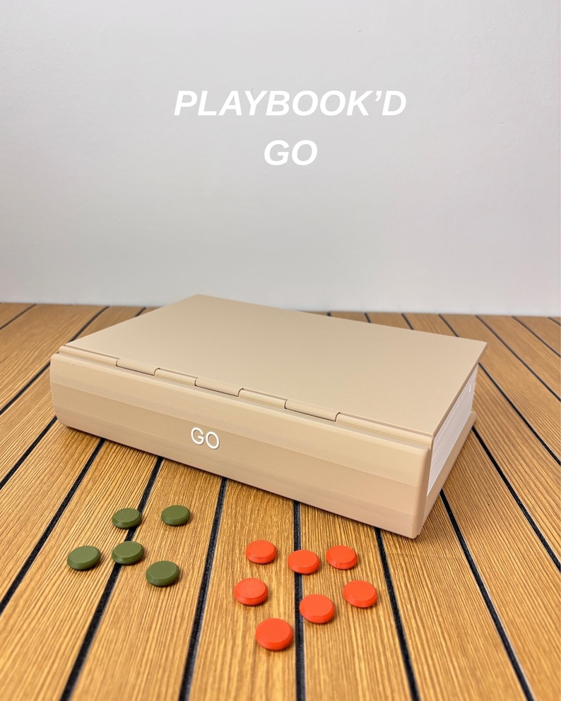 May include: A beige rectangular game case with the text PLAYBOOK'D GO printed above it and the word GO on the front edge. The case is accompanied by four small olive green circular tokens and six small bright orange circular tokens.