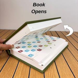 May include: A green hardcover book with the text Book Opens printed above it. The book is partially open, revealing internal pages featuring a grid of white squares with small colored labels containing the text 2L, 3W, and 2W. A white arrow icon points.