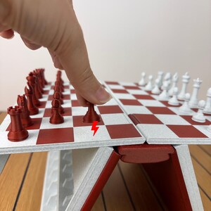 K&ouml;nnte beinhalten: Ein klappbares Schachspiel mit einer rot-wei&szlig; karierten Spielfl&auml;che. Das Set enth&auml;lt rote und wei&szlig;e Schachfiguren, wobei ein roter Bauer auf dem Brett bewegt wird. Ein kleines rotes Blitzsymbol ist auf dem Brett neben dem Bauern aufgedruckt.