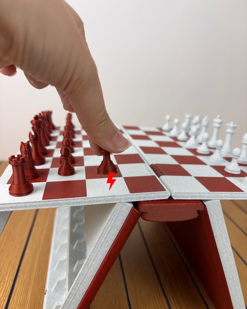 K&ouml;nnte beinhalten: Ein klappbares Schachspiel mit einer rot-wei&szlig; karierten Spielfl&auml;che. Das Set enth&auml;lt rote und wei&szlig;e Schachfiguren, wobei ein roter Bauer auf dem Brett bewegt wird. Ein kleines rotes Blitzsymbol ist auf dem Brett neben dem Bauern aufgedruckt.