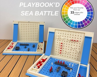 May include: Two folding sea battle board games with cream-coloured frames and blue playing surfaces. Each board features a grid with letters A-J and numbers 1-10, grey plastic ships, and red and white pegs. Text reads PLAYBOOK'D SEA BATTLE.