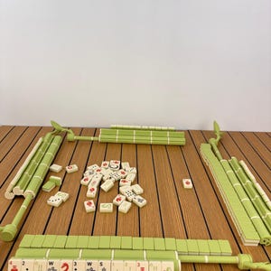 May include: A Mahjong set featuring light green tile racks shaped like bamboo stalks and cream-colored tiles with red, black, and green symbols. The set includes a central pile of loose tiles and four racks arranged in a square formation.