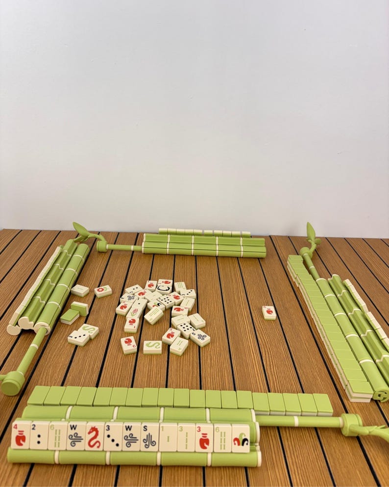 May include: A Mahjong set featuring light green tile racks shaped like bamboo stalks and cream-colored tiles with red, black, and green symbols. The set includes a central pile of loose tiles and four racks arranged in a square formation.