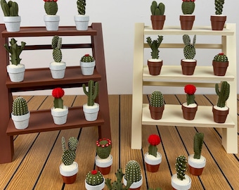 May include: A collection of miniature artificial cactus plants in small pots, displayed on two three-tiered wooden shelves, one dark brown and one cream-coloured. The cacti feature various green shapes, textures, and some have small red flower accents.
