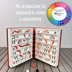Puede incluir: Un juego de mesa de Serpientes y Escaleras dentro de un estuche con forma de libro y cubiertas naranjas. El interior presenta estantes blancos con serpientes naranjas y escaleras verdes. Incluye un dado naranja y tres esferas pequeñas.