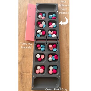 Pode incluir: Um jogo de tabuleiro Mancala cinza com detalhes rosa, cheio de seixos coloridos. O jogo inclui uma capa rosa e o texto "Turns into Mancala" e "Four Different Color Pebbles". O esquema de cores &eacute; rosa e cinza.