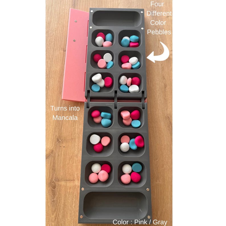 Pode incluir: Um jogo de tabuleiro Mancala cinza com detalhes rosa, cheio de seixos coloridos. O jogo inclui uma capa rosa e o texto "Turns into Mancala" e "Four Different Color Pebbles". O esquema de cores &eacute; rosa e cinza.