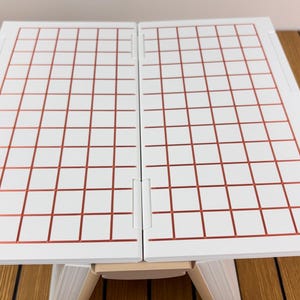 May include: A white folding board featuring a grid pattern of thin, metallic copper-colored lines. The board is composed of two rectangular panels connected by a central hinge, supported by a light-colored base structure underneath.